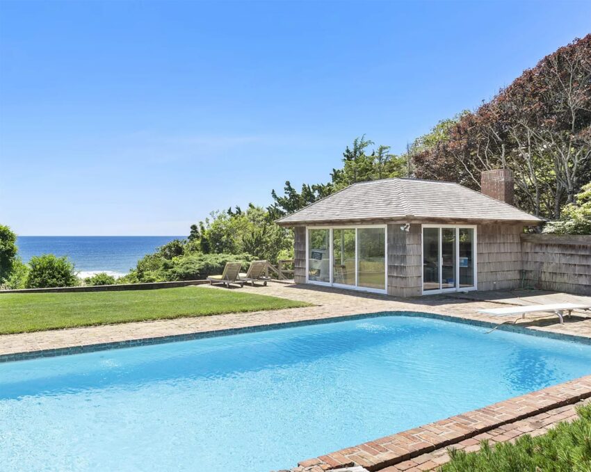 Ocean-view backyard with a swimming pool, cozy pool house, and lush greenery under a clear blue sky.