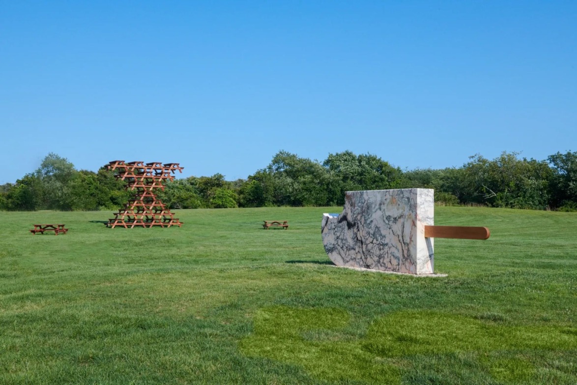 Outdoor sculptures on a grassy field under a clear blue sky with wooden benches and trees in the background