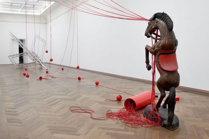 Wooden horse sculpture entangled in red strings with scattered geometric structures in contemporary art gallery space.