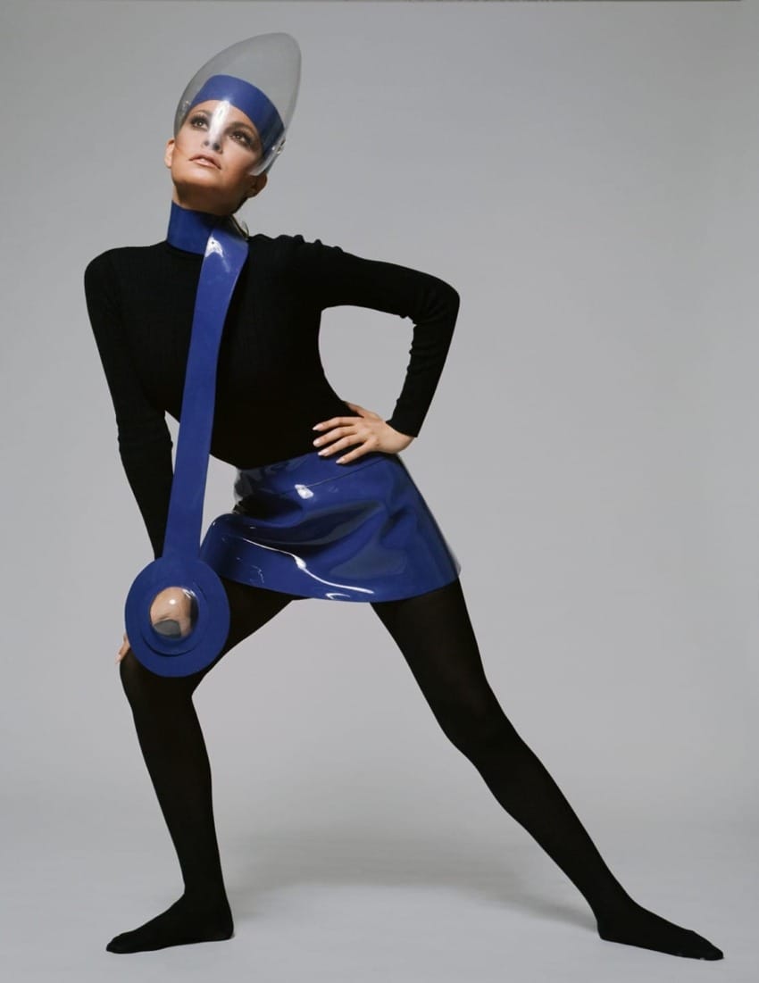 Person posing in a futuristic outfit with a blue visor, black shirt, and blue skirt against a gray background.