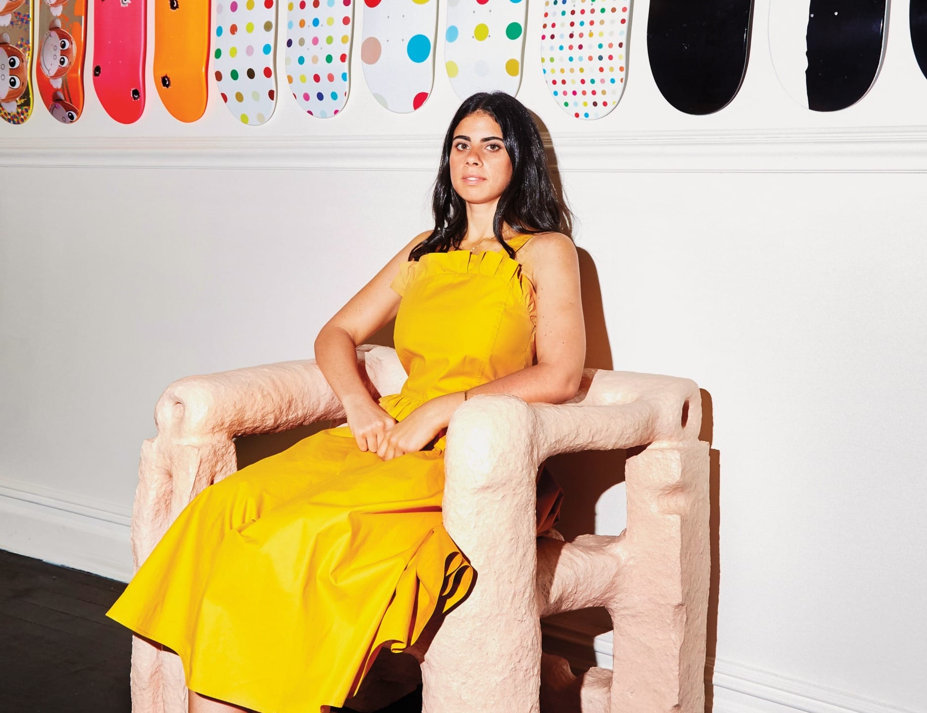 Person in a yellow dress sitting on a uniquely shaped chair with colorful skateboards displayed on the wall behind.