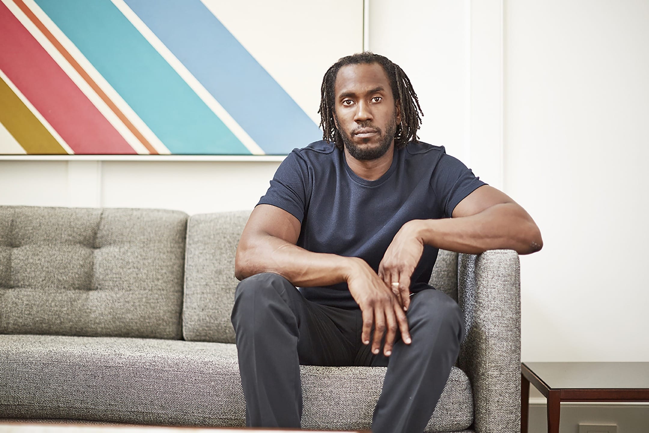 Man sitting on a gray sofa with colorful abstract art in the background, wearing a dark t-shirt and pants.