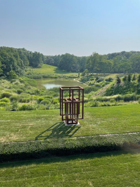 Metal cube sculpture on grass with trees and pond in background under clear blue sky.