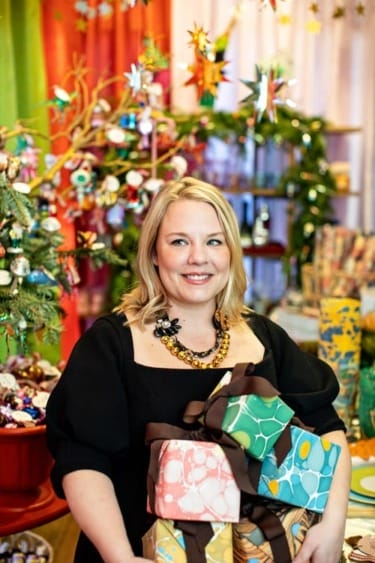 Person holding wrapped gifts in a colorful, festive holiday setting with decorations and lights in the background.