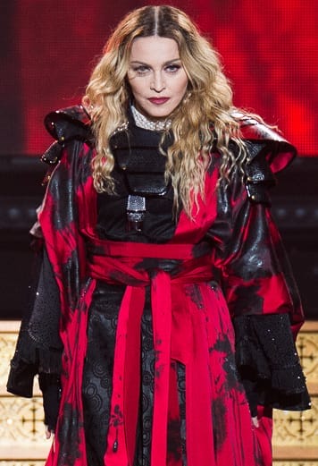 Person in elaborate red and black costume with long wavy hair, performing on stage with a red background.