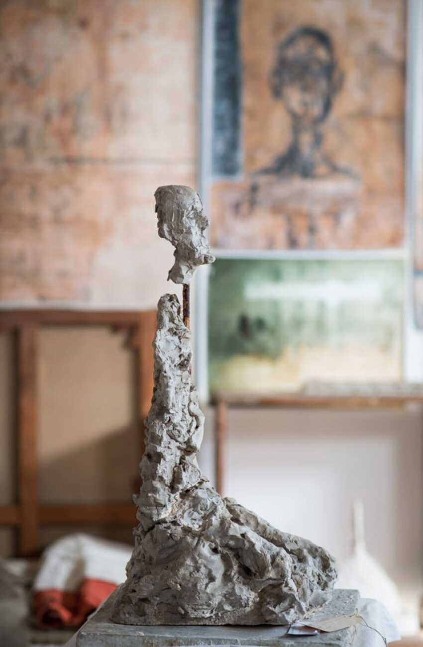 Clay sculpture of a seated figure in an art studio with paintings on the wall in the background