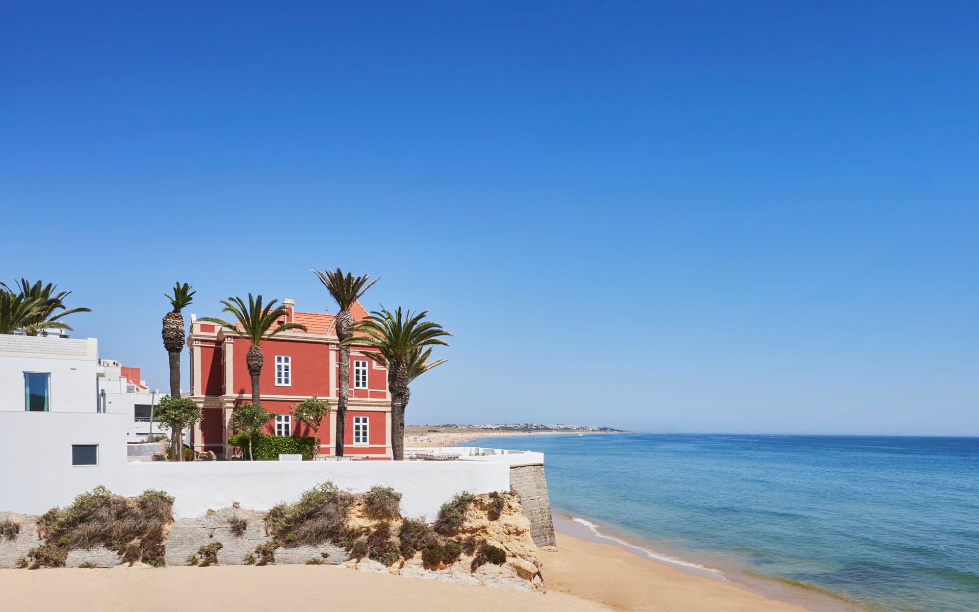 Red house with palm trees on a sandy beach overlooking the clear blue ocean under a bright blue sky.