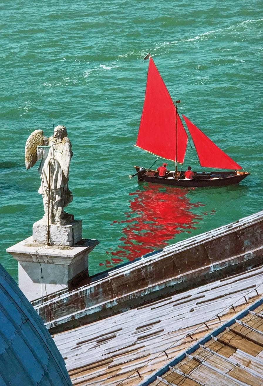 Sailboat with red sails and two people in a canal, seen from a rooftop with angel statue in the foreground.