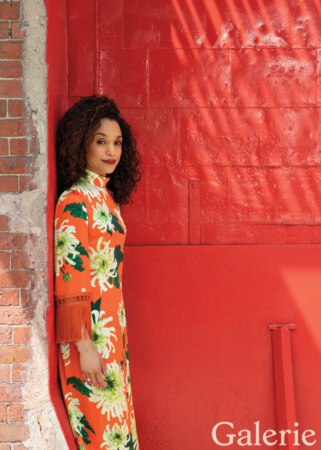 Person in floral dress standing against a vibrant red wall with shadow patterns, Galerie text in the corner.