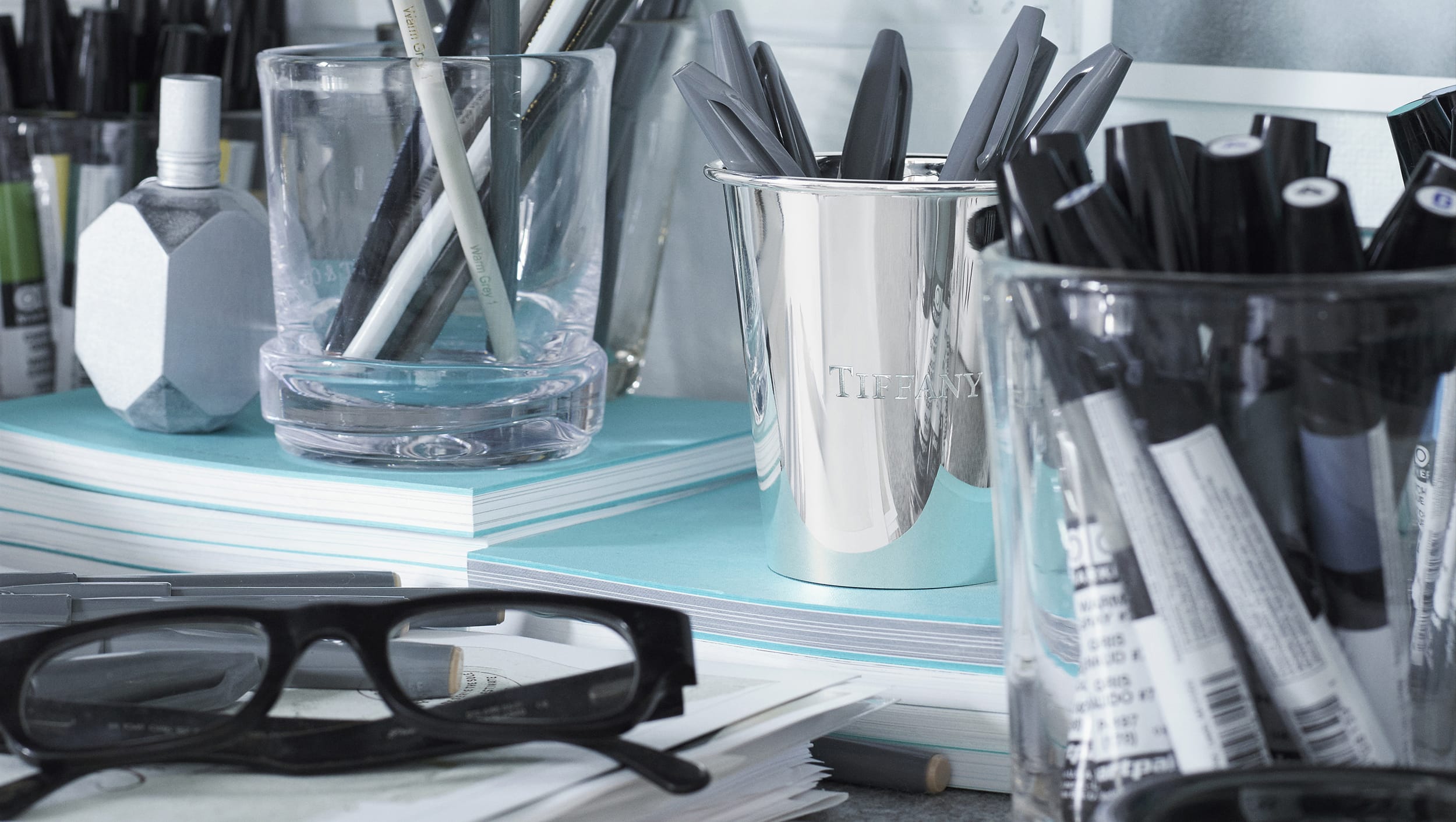 Desk cluttered with glasses, pens, markers in a Tiffany & Co. cup, and notebooks.