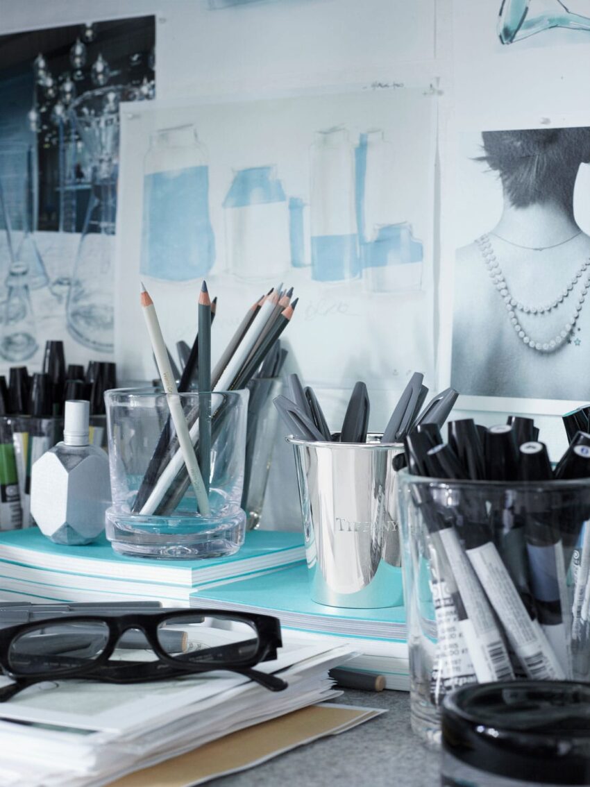 Art studio desk with sketchbooks, pencils, and markers, surrounded by drawings and photos pinned on the wall.
