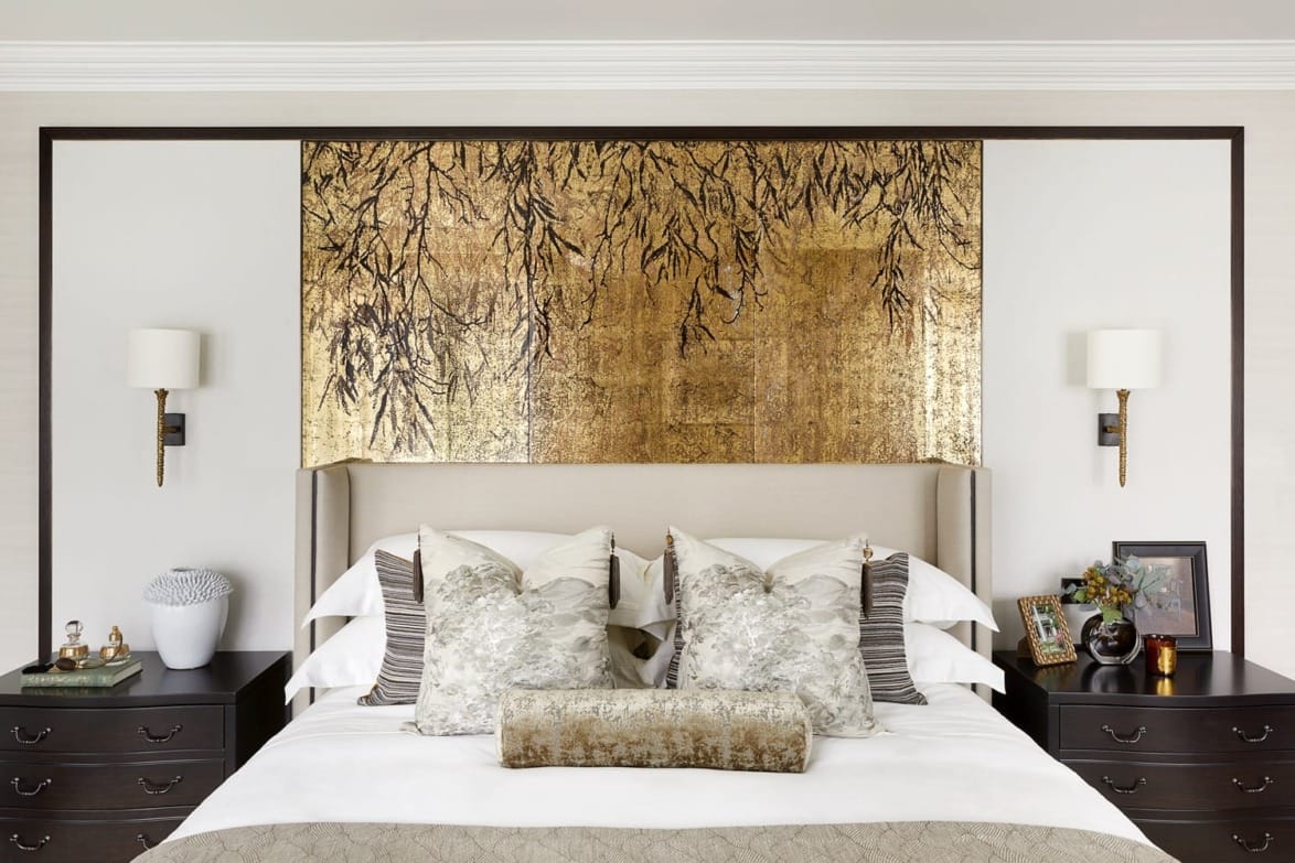 Elegant bedroom with a decorative golden headboard, patterned pillows, matching nightstands, and wall sconces.