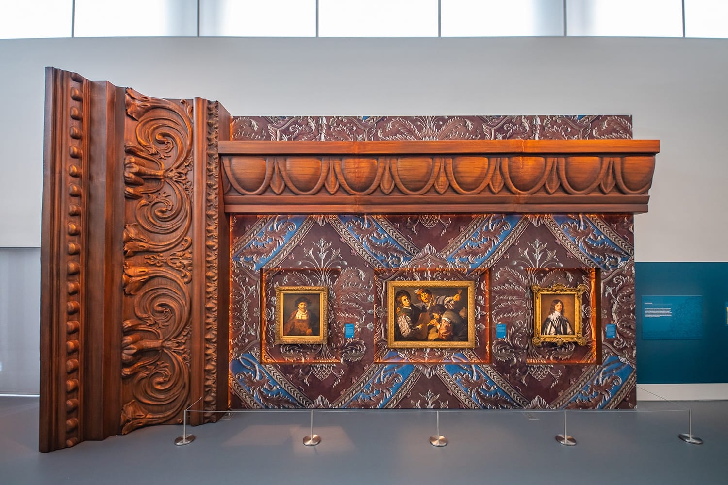 Large, ornate wooden book sculpture with framed paintings embedded on a decorative wall in a modern museum setting.