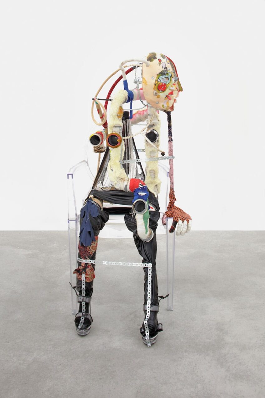 Abstract sculpture of a humanoid figure made from mixed materials, including fabric, metal, and pipes, in a seated position.