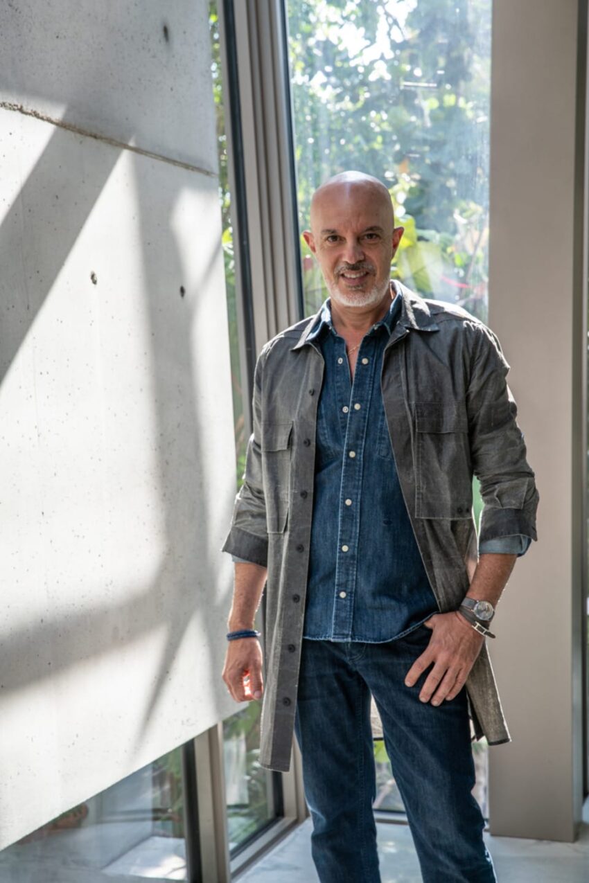 Man in denim outfit standing by large windows with greenery outside.