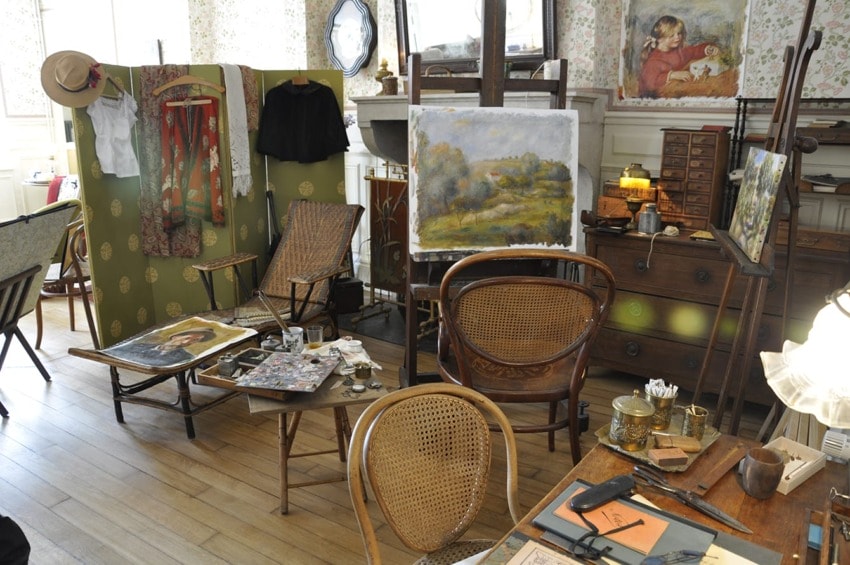 Artist's studio with paintings, easels, chairs, and scattered art supplies, showcasing a creative and cluttered workspace.