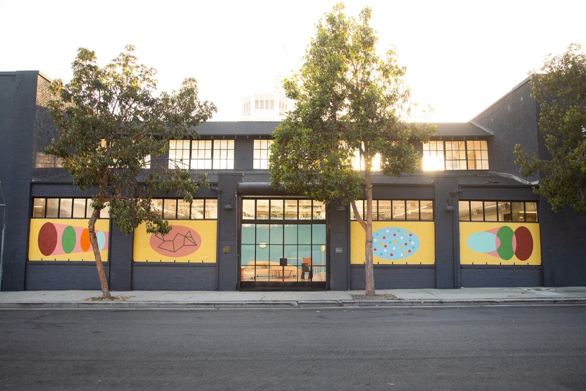A modern industrial building with colorful abstract murals on its facade, flanked by trees along an empty street.