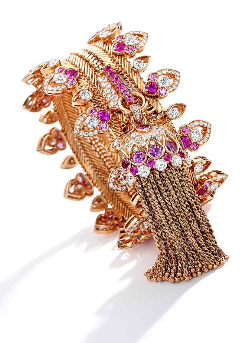 Intricate gold bracelet with floral accents, pink gemstones, and tassel detail on white background.