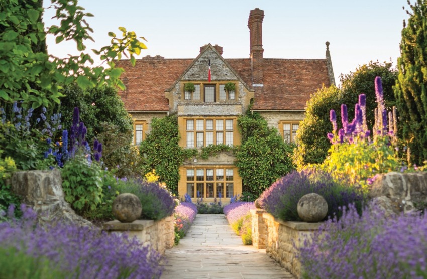 Pathway leading to a charming stone house with a red-tiled roof, surrounded by lush greenery and vibrant purple flowers.