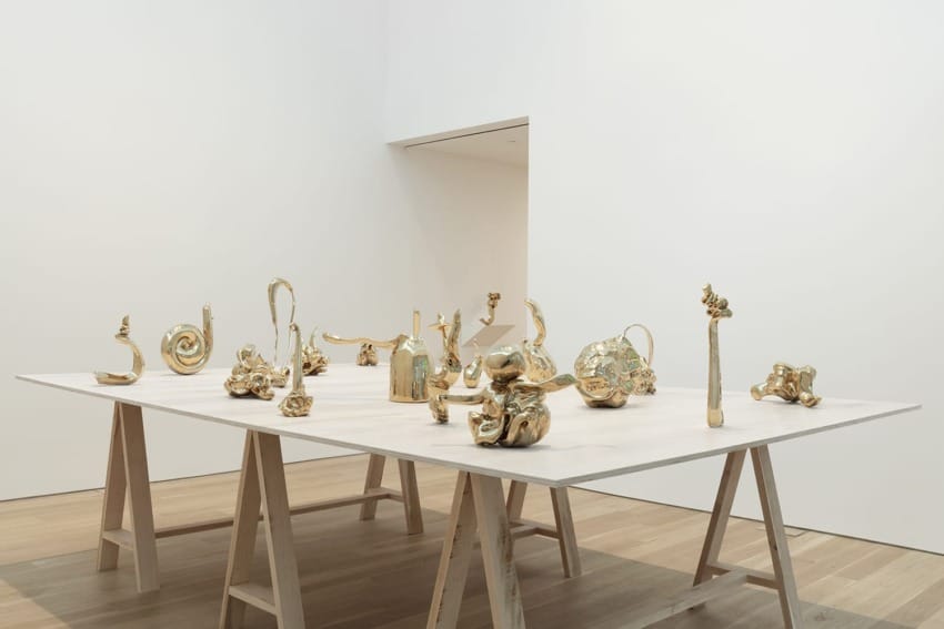 Golden abstract sculptures displayed on a wooden table in a gallery setting with white walls and wooden floors.