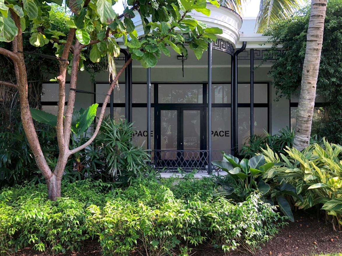 Lush green plants and trees in front of a modern glass building with a black railing and an overhanging roof design.