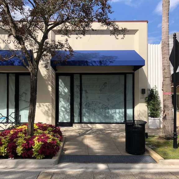 Storefront with blue awning, large windows, and trees; vibrant flowers and a palm tree in front on a sunny day.