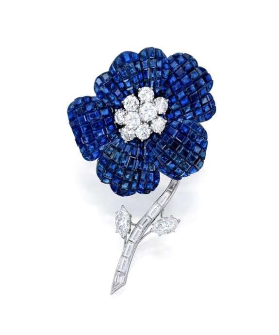 Blue sapphire flower brooch with diamond center and accent on a silver stem against a white background.