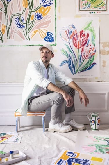 Artist sitting on small chair surrounded by colorful floral artwork on paper, wearing a cap and casual attire.