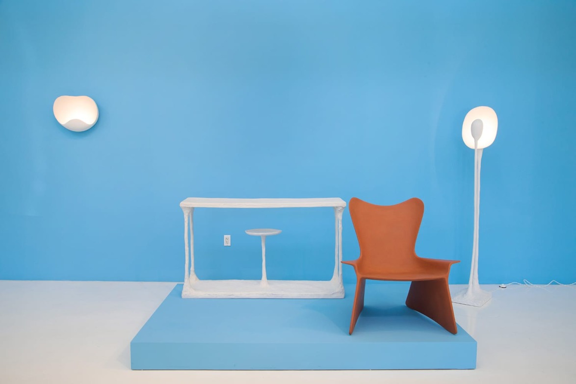 Modern art installation featuring white table, small stool, and brown chair against a bright blue wall with artistic lighting.