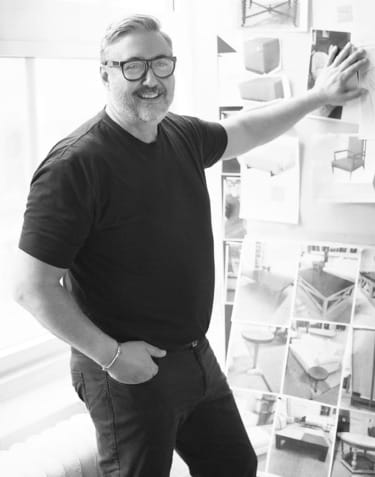 Man smiling and gesturing towards a wall of design sketches and photos in an office setting.