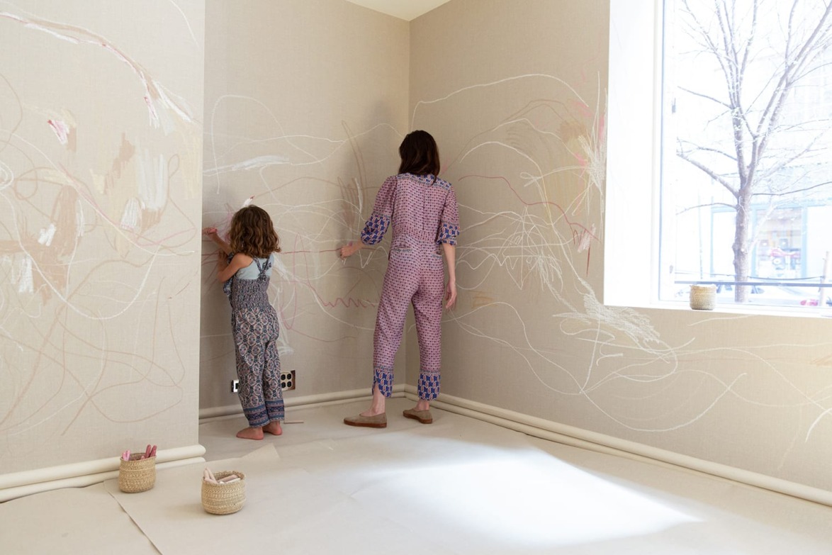 Child and adult drawing on beige walls in a light-filled room with large windows and art supplies nearby.