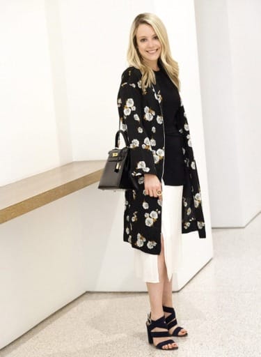 Woman in floral black coat and white skirt stands beside a wooden ledge, holding a black handbag, smiling at the camera.