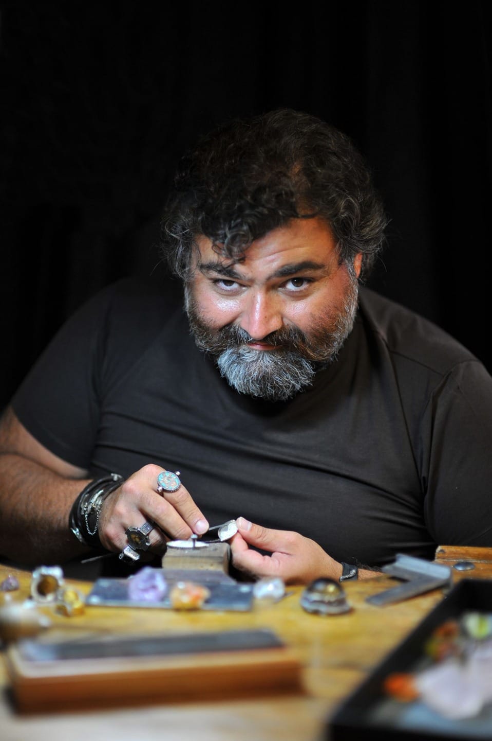 Man working with gemstones at a table, using tools, with various jewelry pieces placed around him.