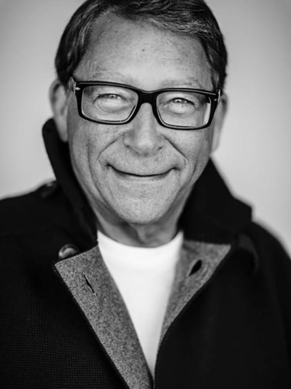 Man wearing glasses and a coat smiling in a black and white portrait.