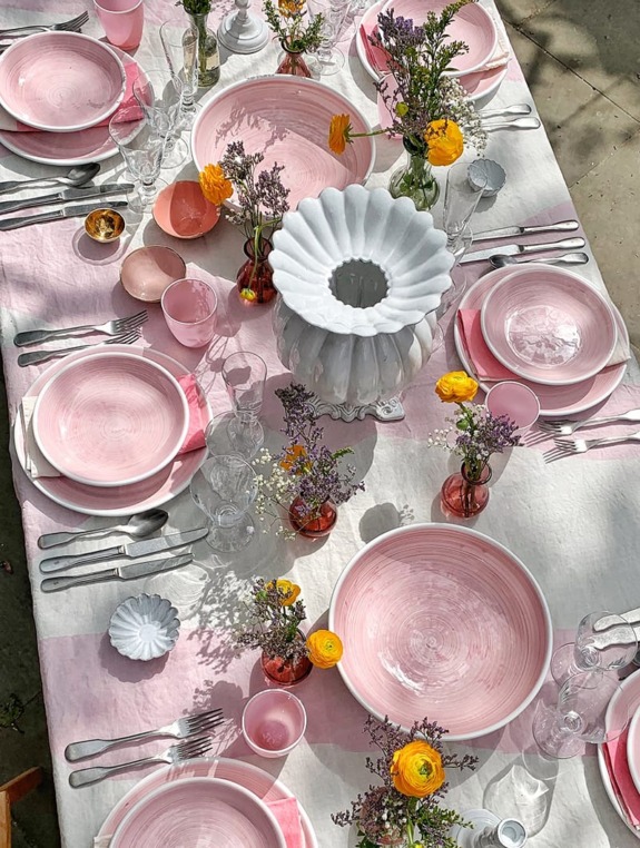 Elegant table setting with pink and white dishes, colorful flowers, and a white vase on a pink tablecloth.