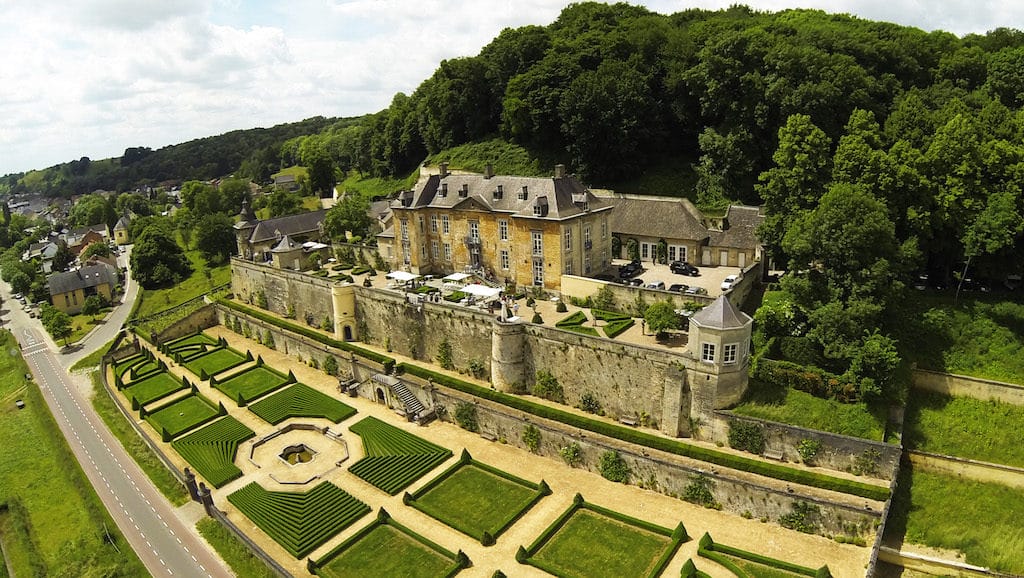 Aerial view of a historic chateau surrounded by manicured gardens and lush greenery on a sunny day.