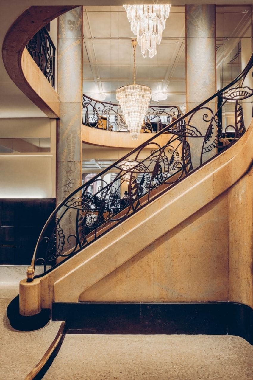 Elegant spiral staircase with ornate railing and two chandeliers in a luxurious interior setting.