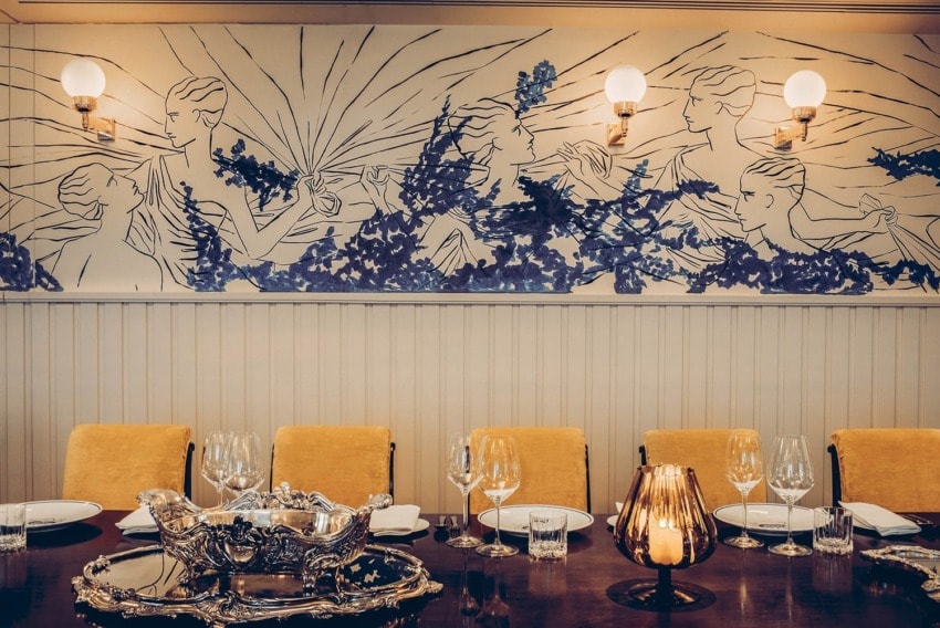 Elegant dining room with yellow chairs, artistic wall mural, ornate silverware, and neatly set table with glasses and plates.