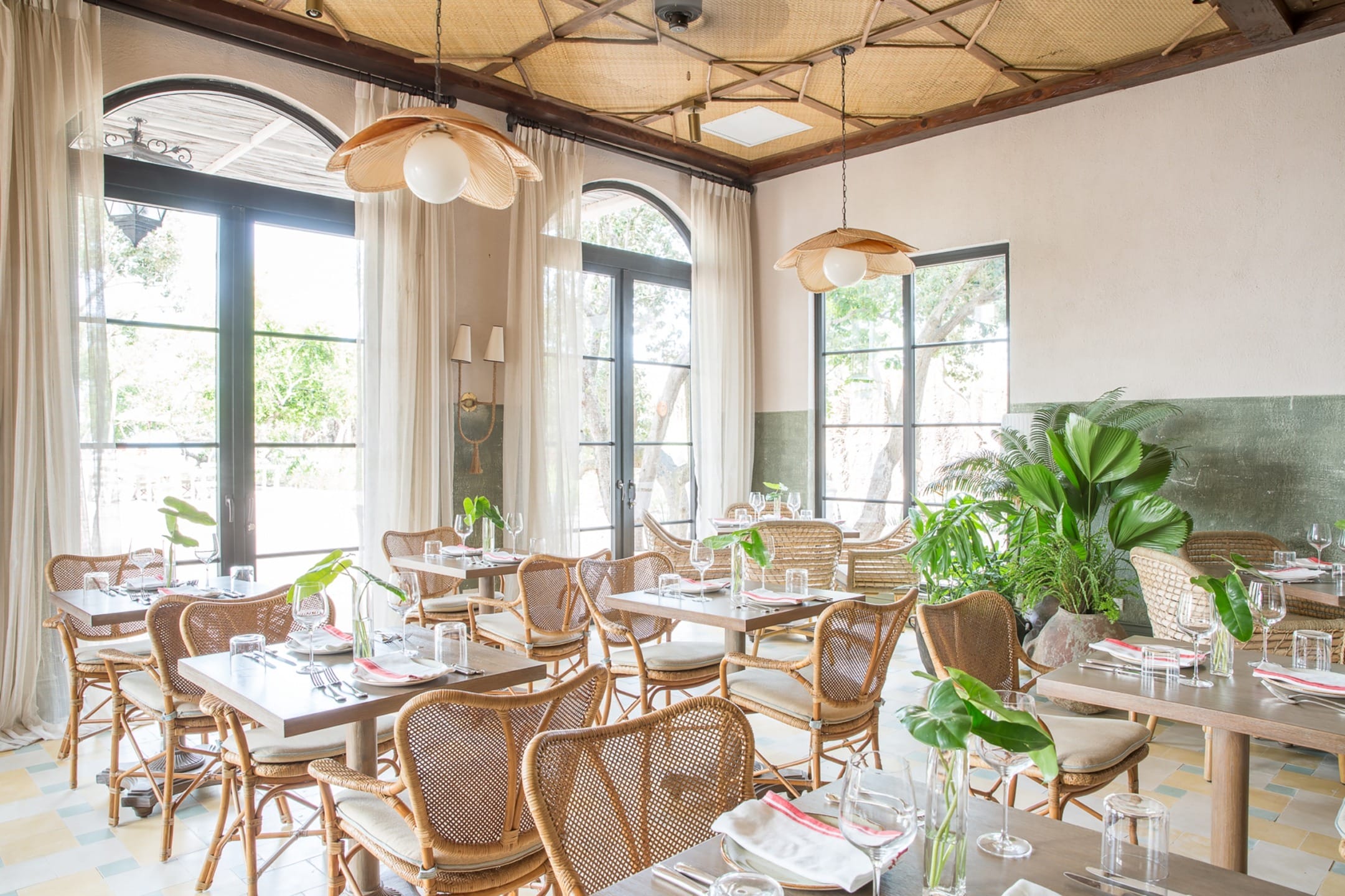 Bright restaurant interior with wicker chairs, wooden tables, and large windows overlooking greenery.