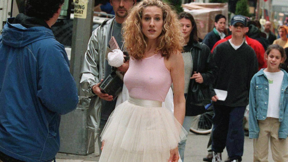 Person in a pink top and white skirt walking on a bustling city street, surrounded by people.
