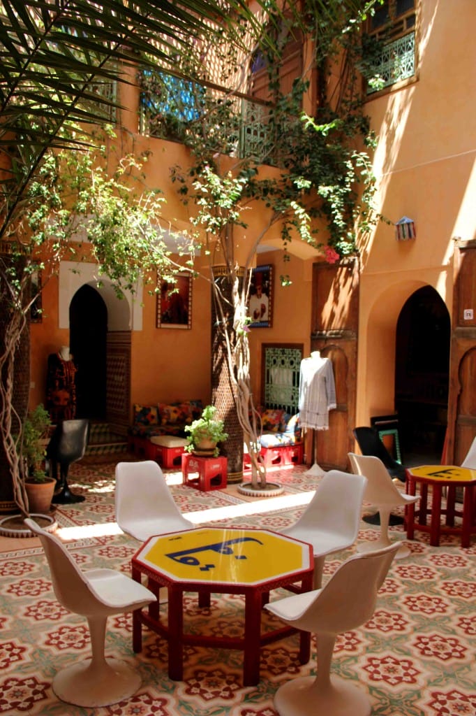 Moroccan courtyard with patterned tiles, round tables, white chairs, and lush greenery under sunlight.