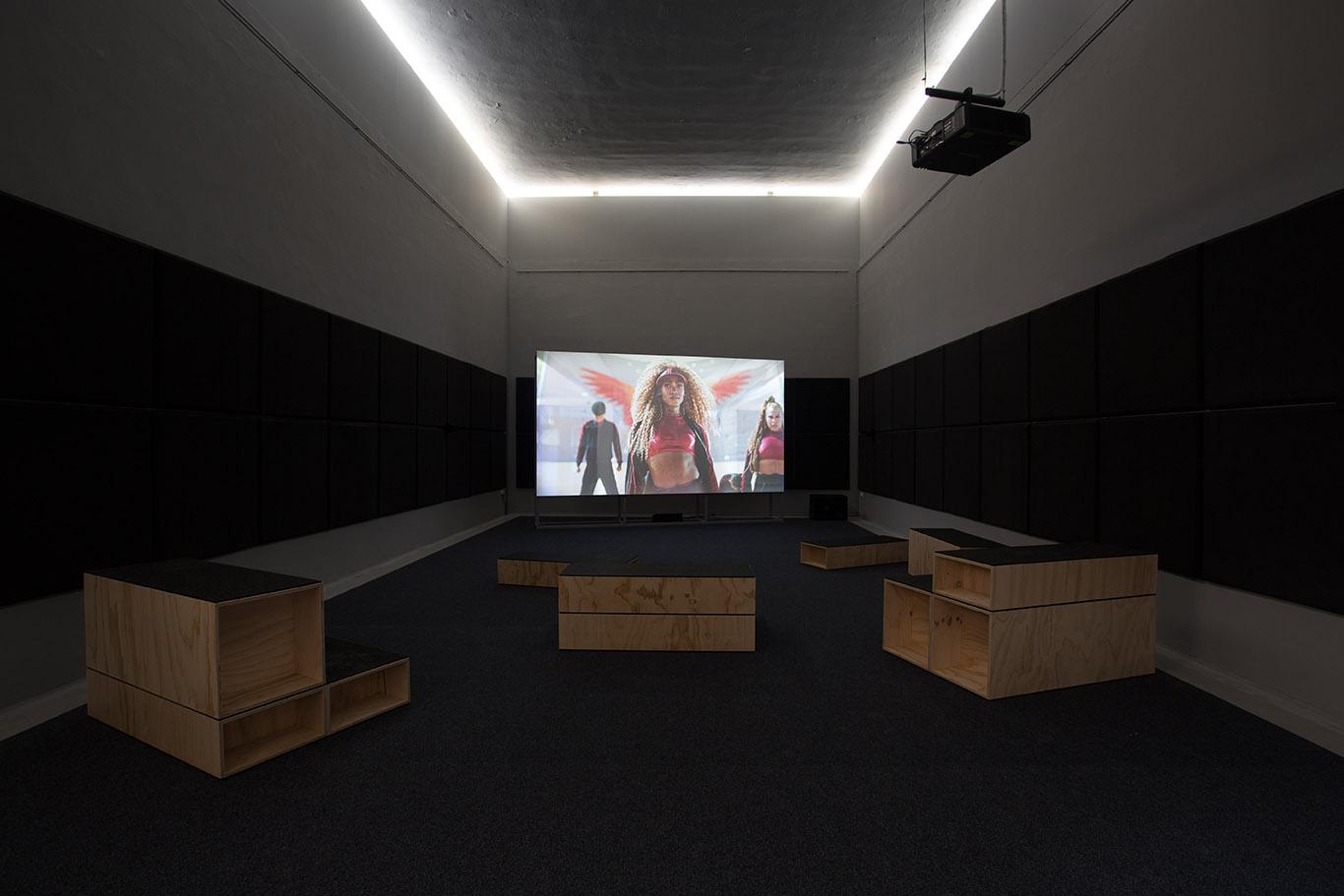 Dark room set up for a film screening with wooden seating and a projector showing a scene with two people.