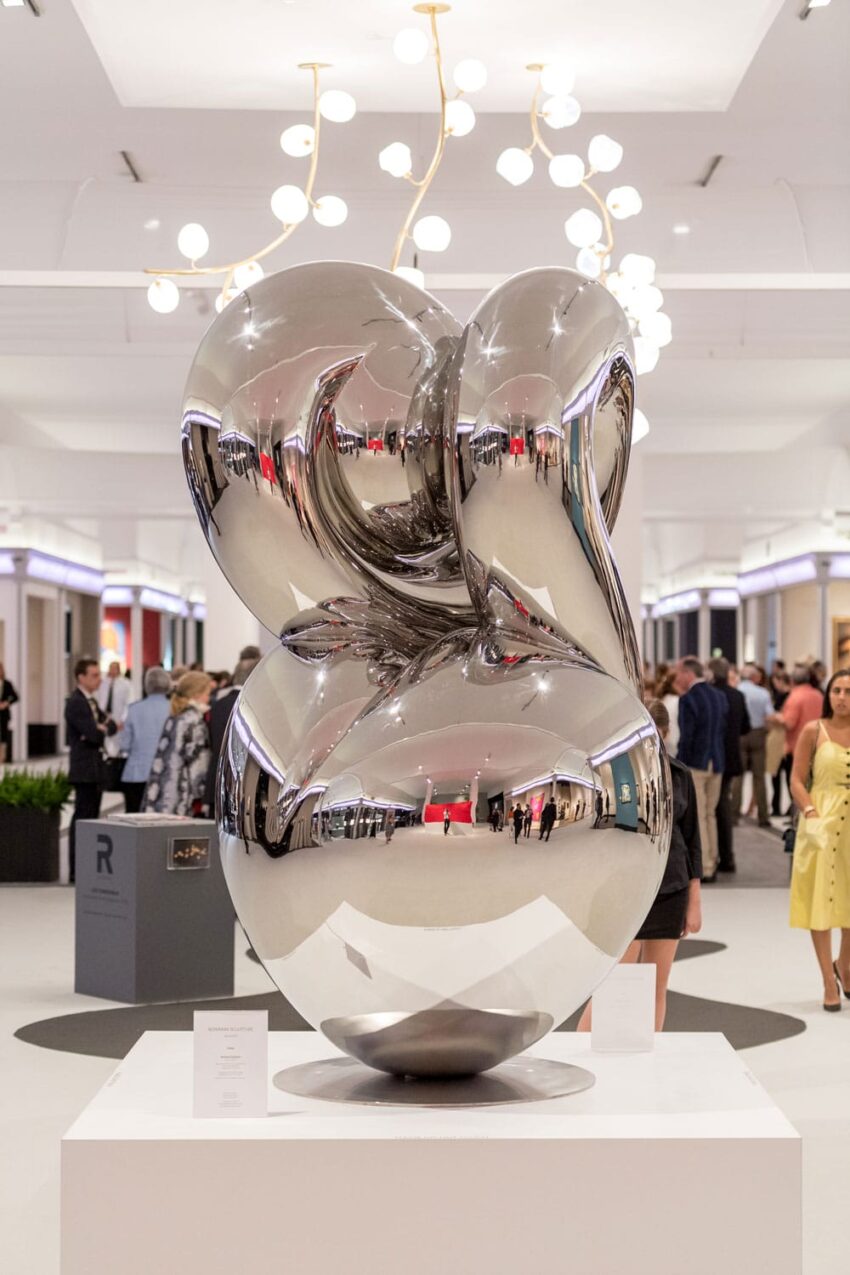Abstract chrome sculpture displayed in a modern exhibition hall with people observing artwork in the background.