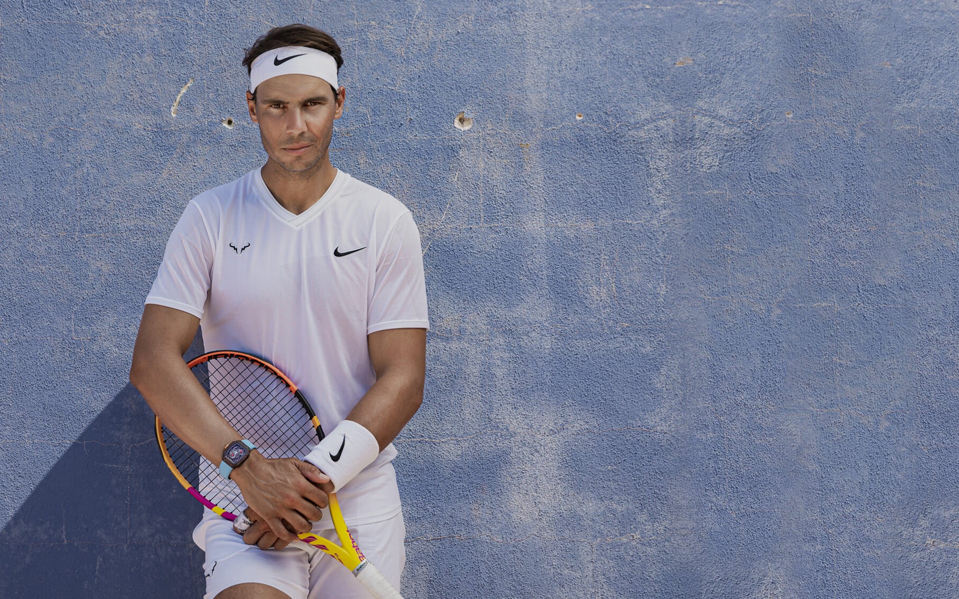 Tennis player in white outfit holding a racket, leaning against a blue wall.
