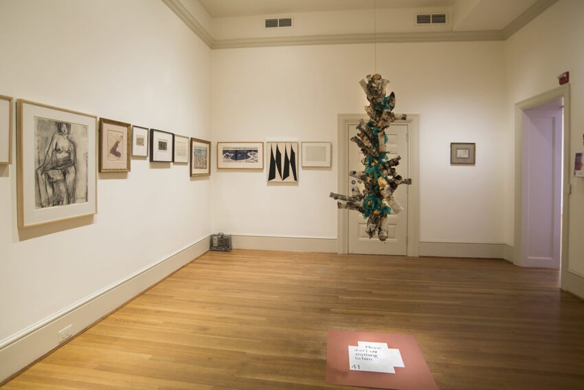 Art gallery interior with framed artwork on white walls and a modern hanging sculpture in the center of the room.