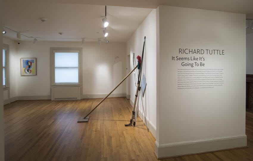 Art gallery interior with wooden floors, abstract wall art, and a metal sculpture leaning against a wall with exhibit details.