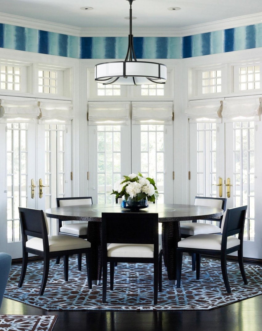 Elegant dining room with round table, black chairs, floral centerpiece, geometric rug, and large windows in the background.
