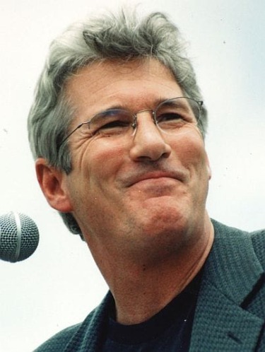 Smiling man with glasses and gray hair, standing near a microphone against a cloudy sky background.