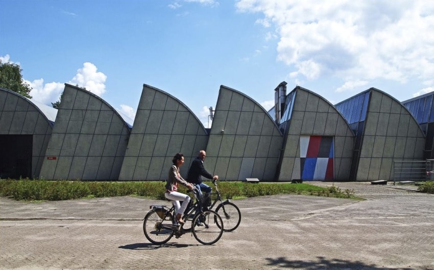 Two people riding bicycles in front of a modern building with curved roofs on a sunny day.