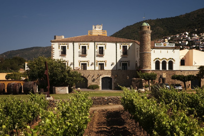 Historic winery with vineyard in foreground, located at the base of a mountain on a sunny day.